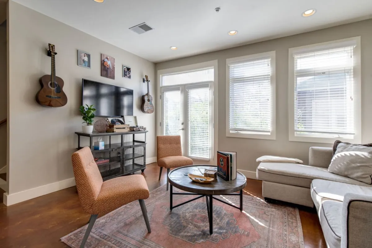 A living room scene with guitars mounted on the wall, a television unit, a round coffee table, and a window letting in abundant natural light. The setup suggests relaxing with friends, watching TV, or enjoying a coffee, and the décor adds a creative, stylish touch.
