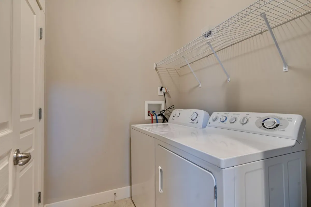 The image displays an in-unit laundry area with a washing machine and dryer stacked neatly, complemented by a shelf above for organizing laundry supplies. The presentation is practical and tidy, highlighting the condo’s ability to support extended stays with convenient amenities.