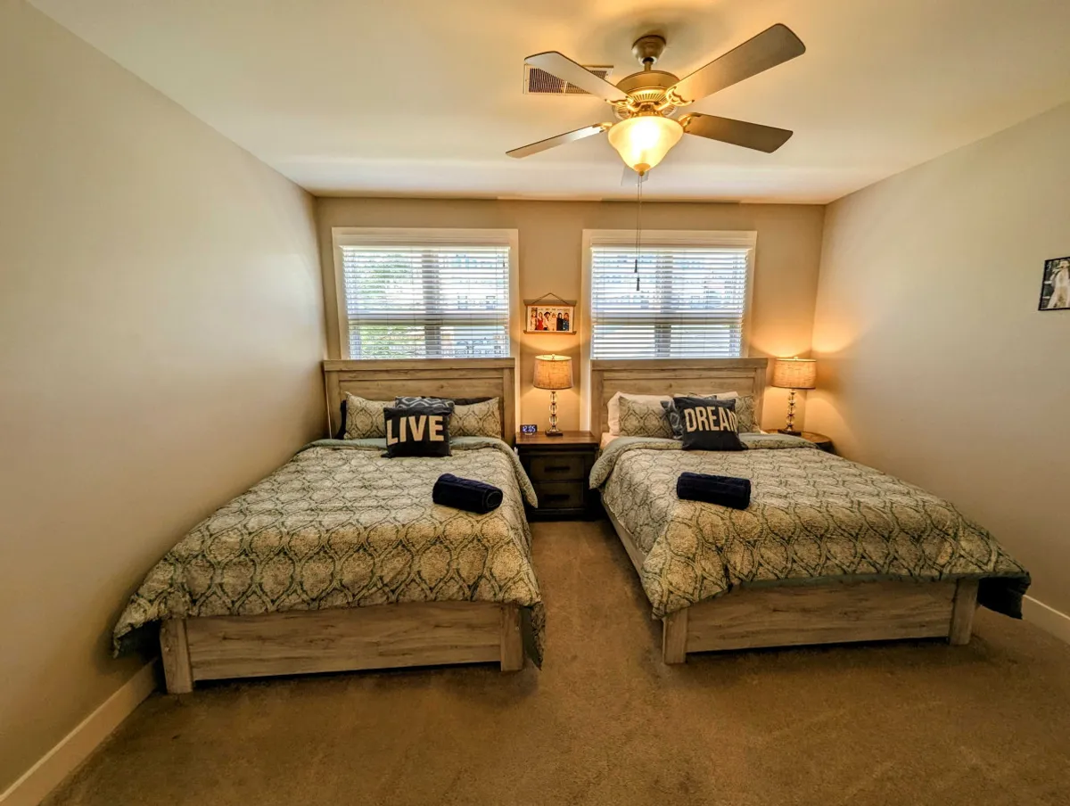 This image captures a comfortable bedroom with two beds adorned with decorative pillows, nightstands with lamps on either side, and windows with blinds, allowing natural light to softly illuminate the space. The overall atmosphere is warm and inviting, designed for relaxation and socializing.
