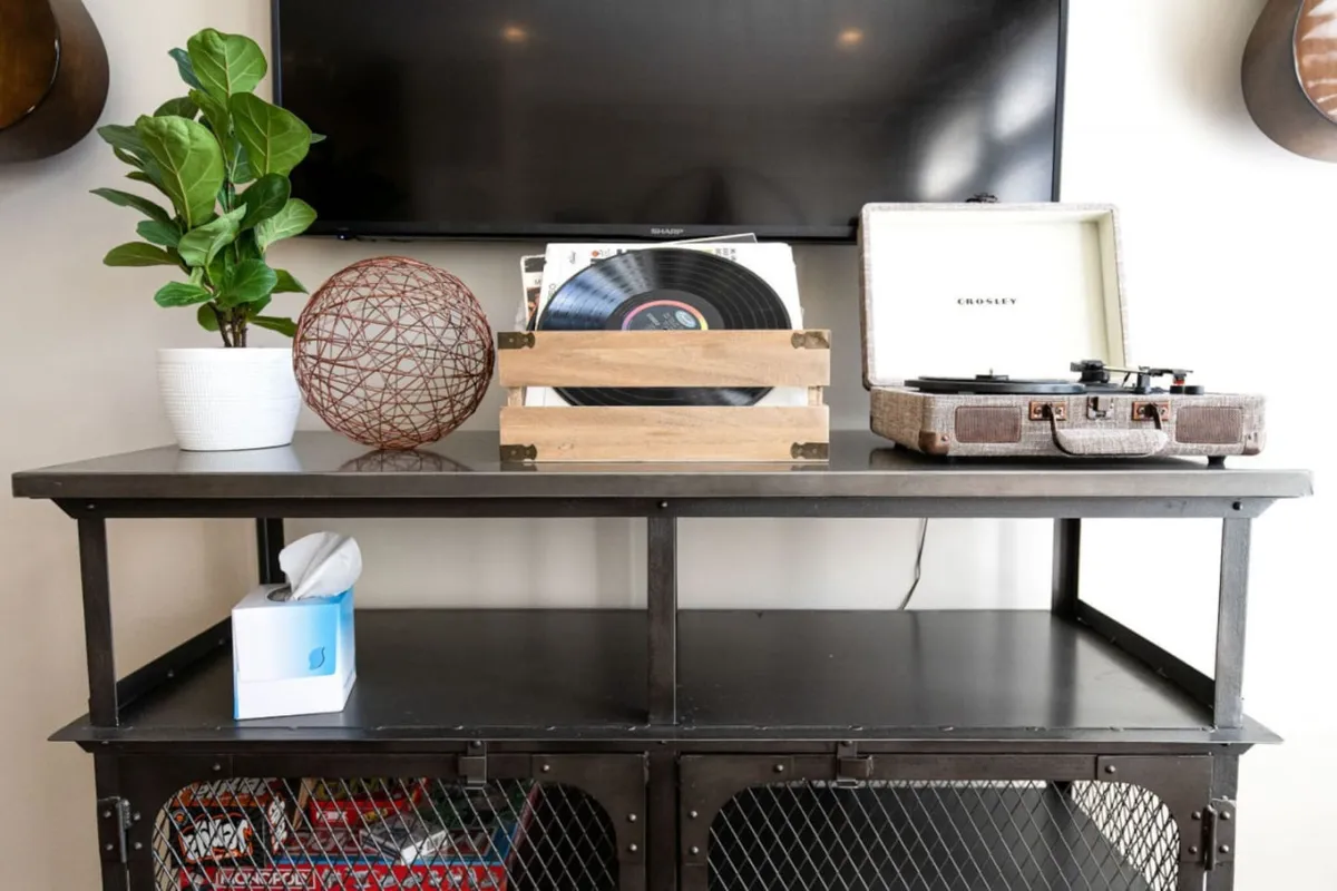 The image features a cozy interior scene centered around a classic record player surrounded by vinyl records, a decorative plant, and a woven sphere. The mood is nostalgic and warm, with suggestions of guests sharing music and stories, relaxing together, and enjoying each other's company in an inviting living space.