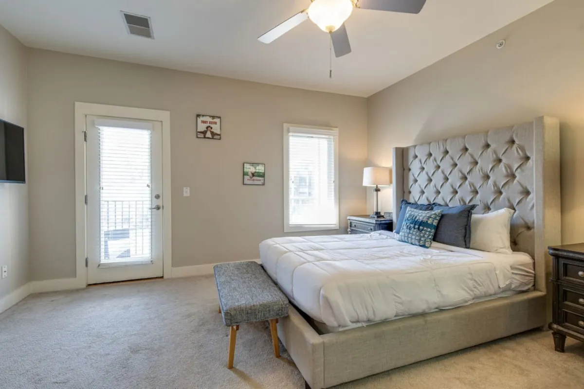 A serene bedroom featuring a king-sized bed dressed with decorative pillows, a modern bedside lamp, a stylish bench at the foot of the bed, framed artwork, and a large window that bathes the room in daylight. The setting communicates comfort and tranquility, ideal for restful moments and private escapes.